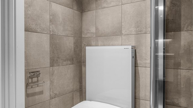 Badet er utstyrt med vegghengt toalett og dusjhjørne med innfellbare glassdører.