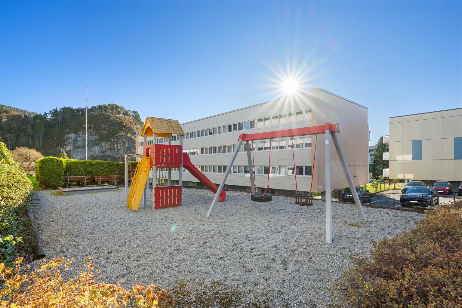 Skjermet lekeplass med husker og lekestativ like utenfor.