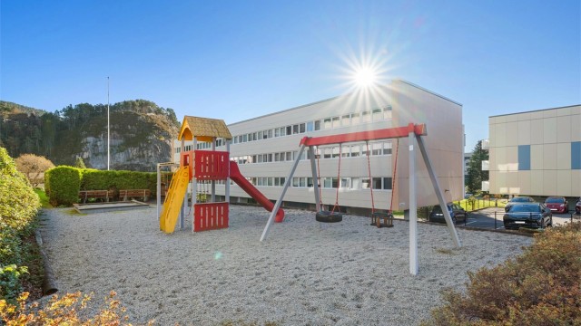 Skjermet lekeplass med husker og lekestativ like utenfor.