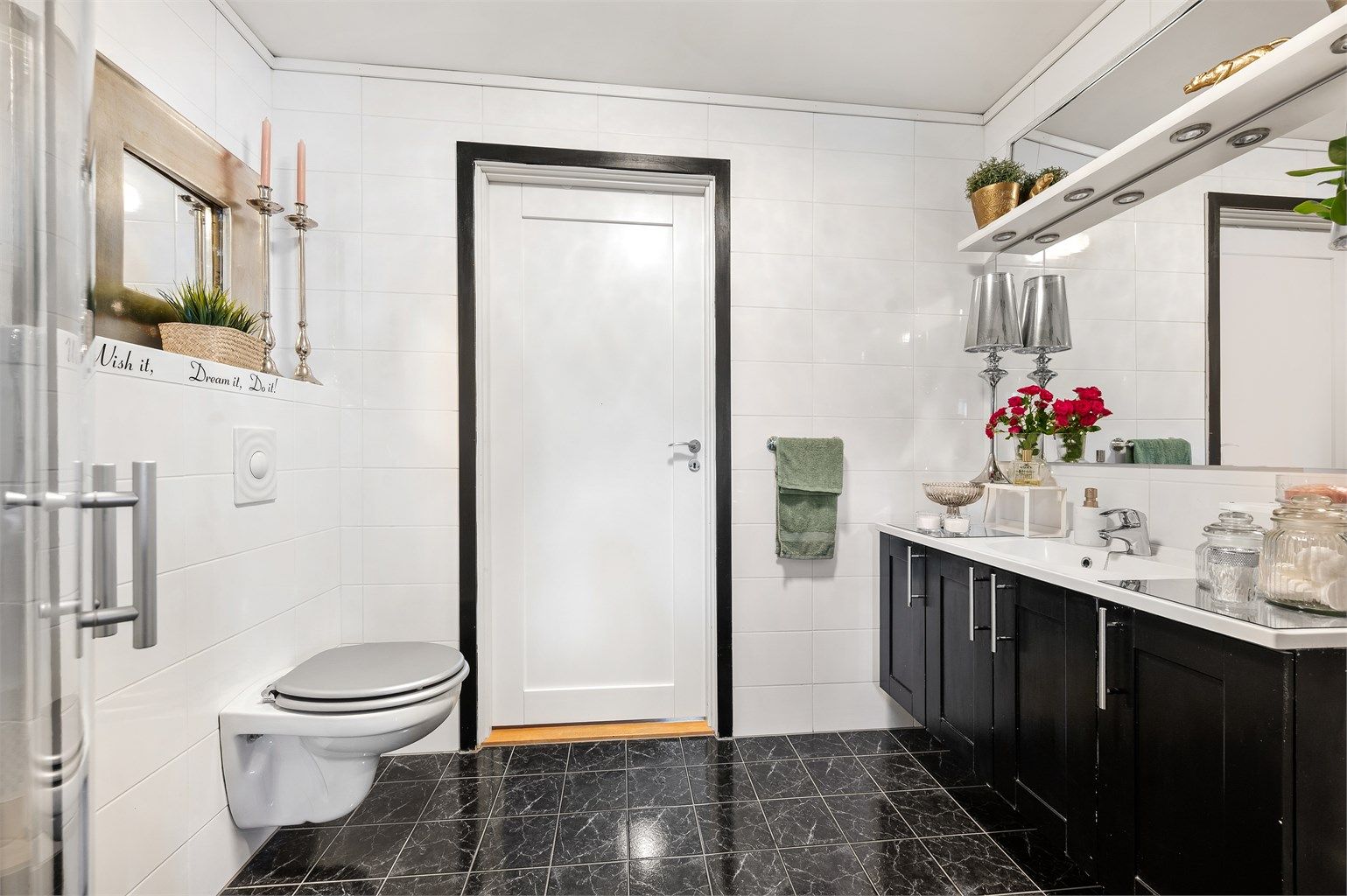 Badet i første etasje er innredet med mørke gulvfliser med hvite veggfliser, dusjhjørne, servant i innredning, vegghengt toalett og opplegg for vaskemaskin.