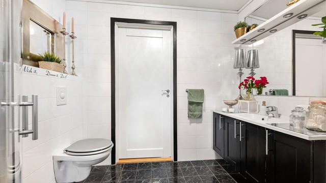 Badet i første etasje er innredet med mørke gulvfliser med hvite veggfliser, dusjhjørne, servant i innredning, vegghengt toalett og opplegg for vaskemaskin.