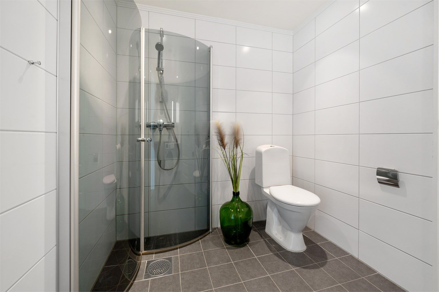 Badet har dusjhjørne med innfellbare glassdører og dusjarmatur, samt gulvmontert toalett.