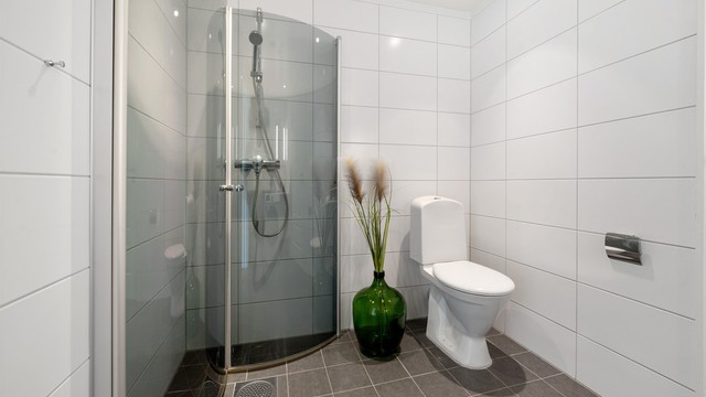 Badet har dusjhjørne med innfellbare glassdører og dusjarmatur, samt gulvmontert toalett.