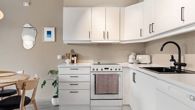 Kjøkkenvask med armatur. Frittstående komfyr, oppvaskmaskin og kjøleskap med fryser. Ventilator integrert i overskap, tilknyttet boligens mekaniske avtrekksventilasjon. Varmtvannsbereder er praktisk plassert under kjøkkenbenken.