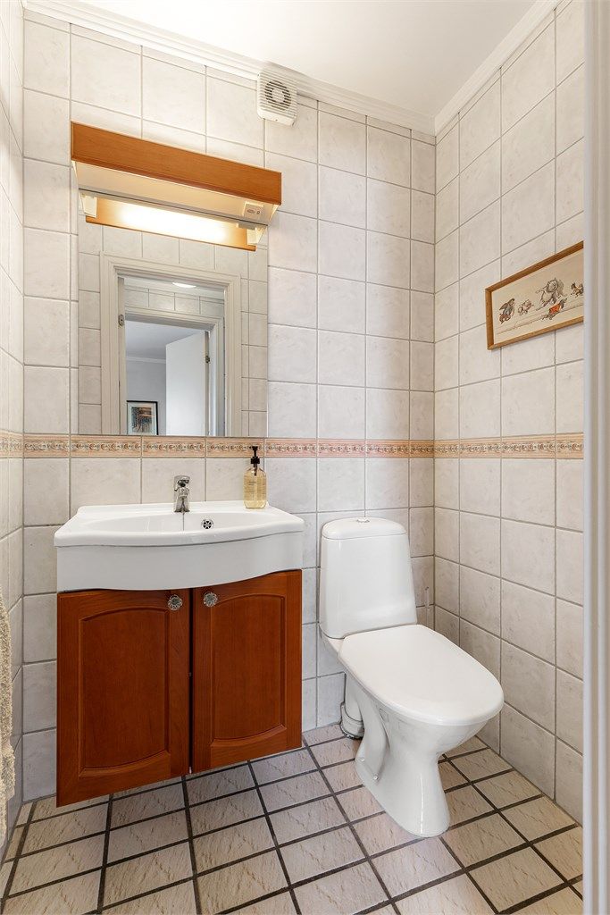 Toalettrom med gulvflate belagt med fliser og gulvvarme. Flislagte vegger. Malte flater i himling. Vegghengt servantinnredning. Ovenpåliggende servant med armatur. Gulvstående toalett.