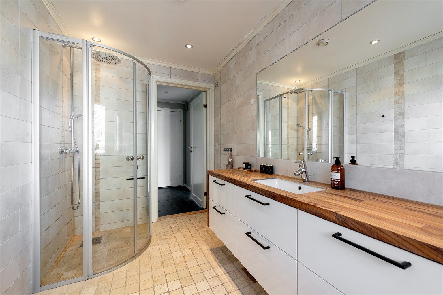 Flislagt bad med gulvvarme. Stor baderomsinnredning med skuffer og nedfelt servant med ett-greps armatur. Stort innfelt speil over servant. Dusjhjørne med dører og vegghengt dusjarmatur med hånddusj og regndusj). Vegghengt toalett. 