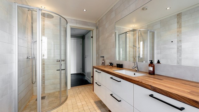 Flislagt bad med gulvvarme. Stor baderomsinnredning med skuffer og nedfelt servant med ett-greps armatur. Stort innfelt speil over servant. Dusjhjørne med dører og vegghengt dusjarmatur med hånddusj og regndusj). Vegghengt toalett.