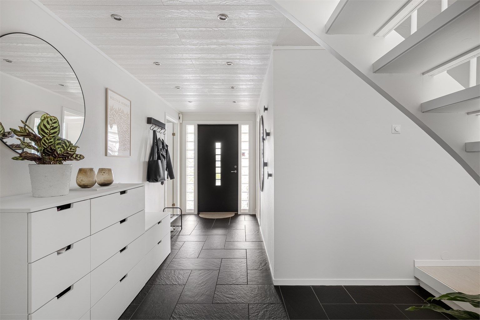 Romslig gang med fliser på gulv og varmekabler. Rikelig med plass til å sette av sko og henge av yttertøy. Spotter i tak. 
