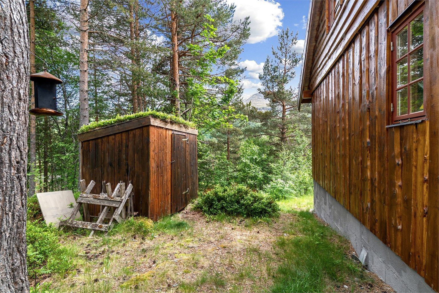 Frittstående vedskjul i trekonstruksjon med torvtak på tomten.
