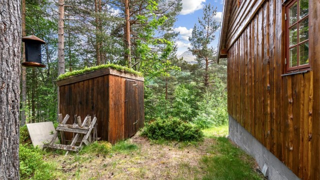 Frittstående vedskjul i trekonstruksjon med torvtak på tomten.