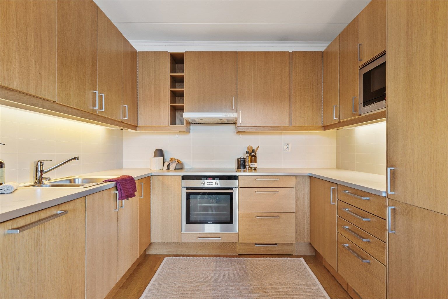 Åpen kjøkkenløsning. Innredning fra byggeår. Glatte fronter. Benkeplate med laminert overflate. Kjøkkenvask med armatur. Stikkontakt over kjøkkenbenk. Integrert stekeovn, platetopp, oppvaskmaskin og kjøleskap med fryser.