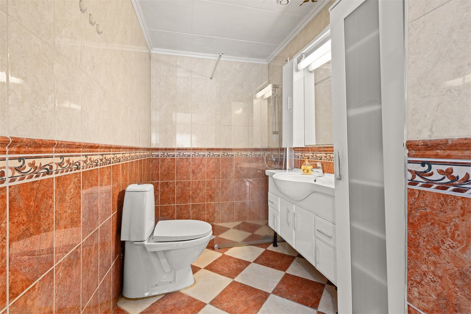 Badet er utstyrt med gulvstående toalett, servantinnredning og dusjnisje med glassvegg.