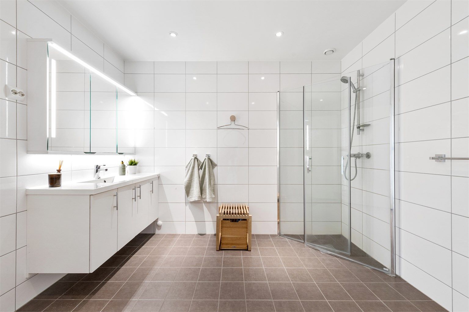 Flislagt bad med gulvvarme. Romsling baderomsinnredning med hvite glatte fronter og ovenpåliggende servant. Speilskap med lys over servant.