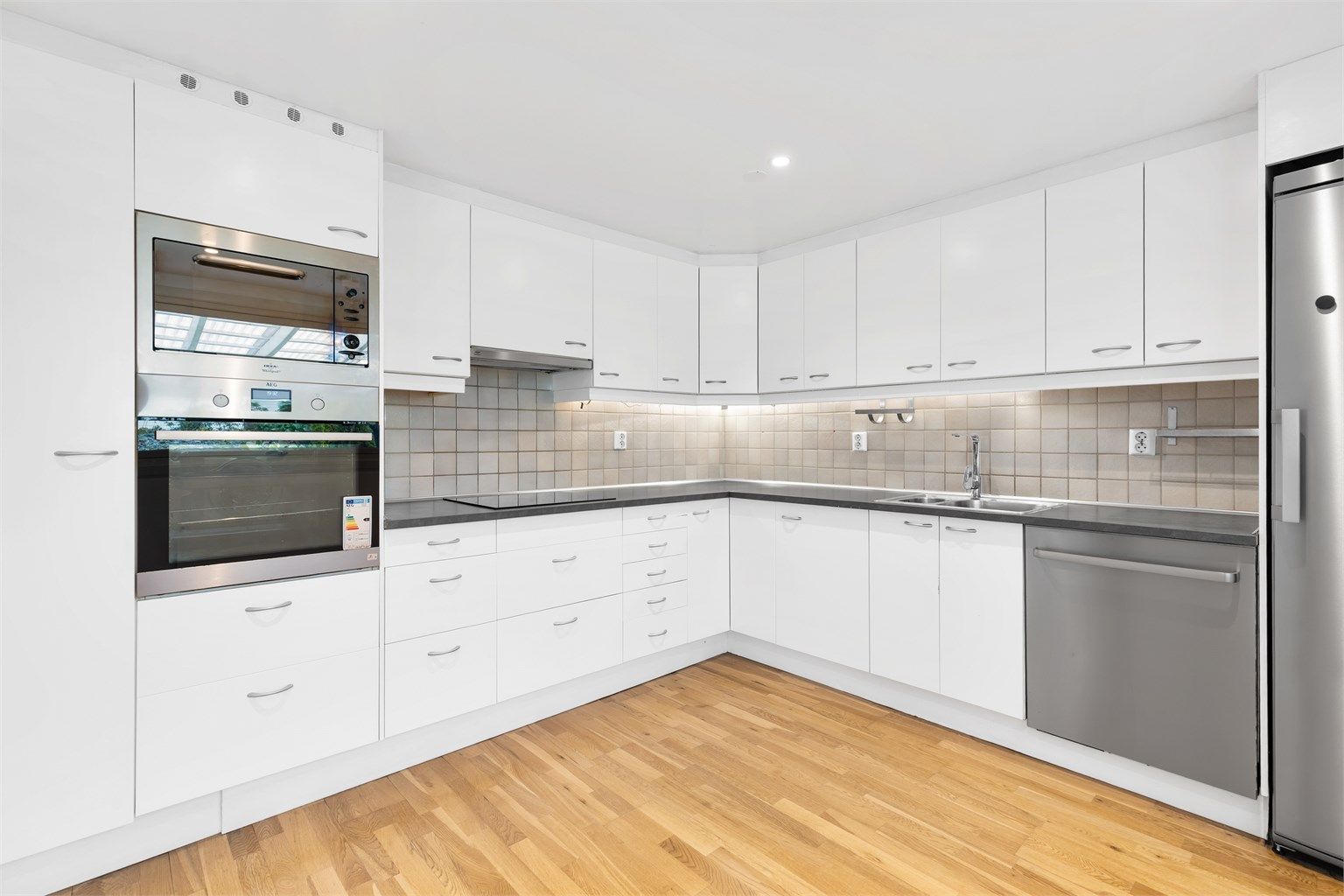 Kjøkkeninnredning med glatte fronter og laminert benkeplate med nedfelt utslagsvask. Det er frittstående oppvaskmaskin og kjøleskap. Integrert komfyr/platetopp og mikrobølgeovn. Mellom over- og underskap er det flislagt.