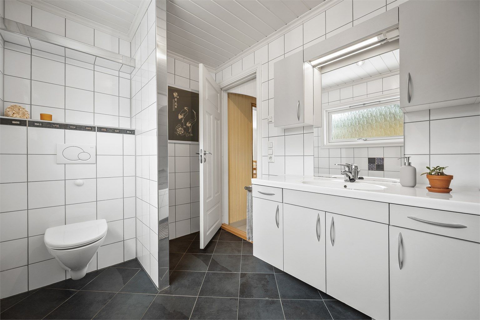 Badet er utstyrt med vegghengt toalett og innfliset badekar med dusjvegg av glass samt veggmontert dusjarmatur