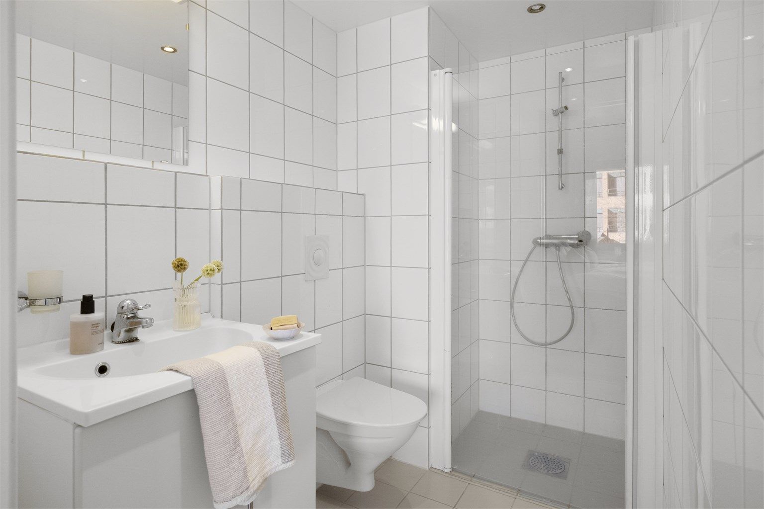 Leilighetens andre bad er også helfliset og har en praktisk dusjløsning. Badet er utstyrt med servantinnredning, speil med belysning og vegghengt toalett.
