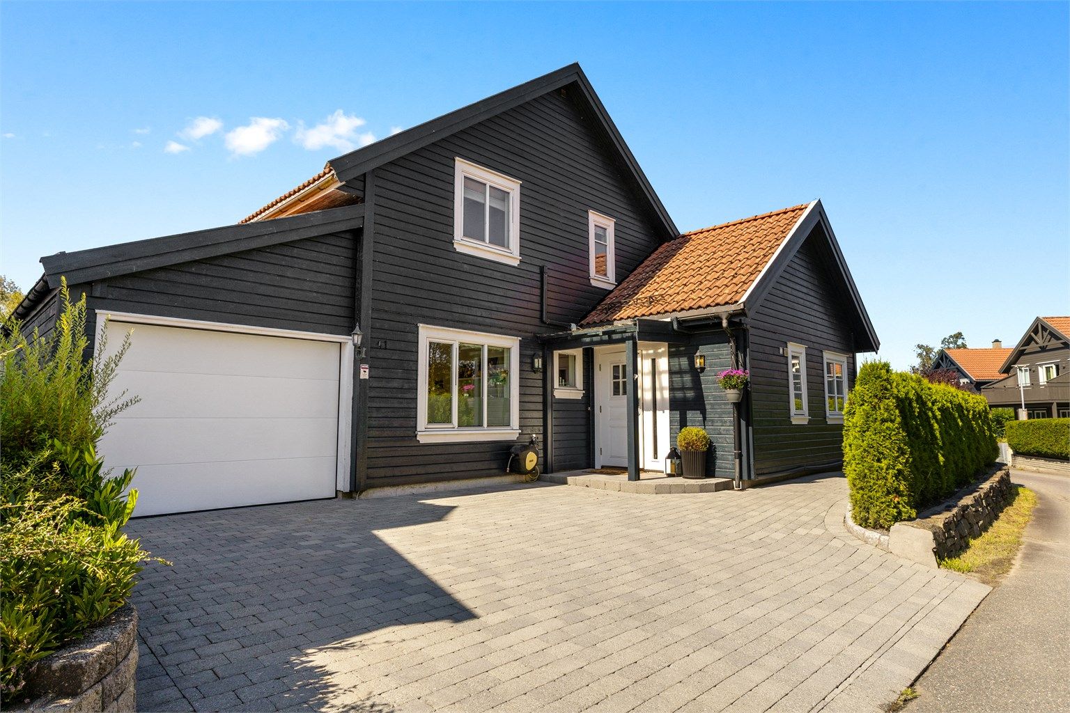 Pent inngangsparti med belegningsstein og beplantning. Garasje med el-bil lader samt parkering til 2 biler utenfor.