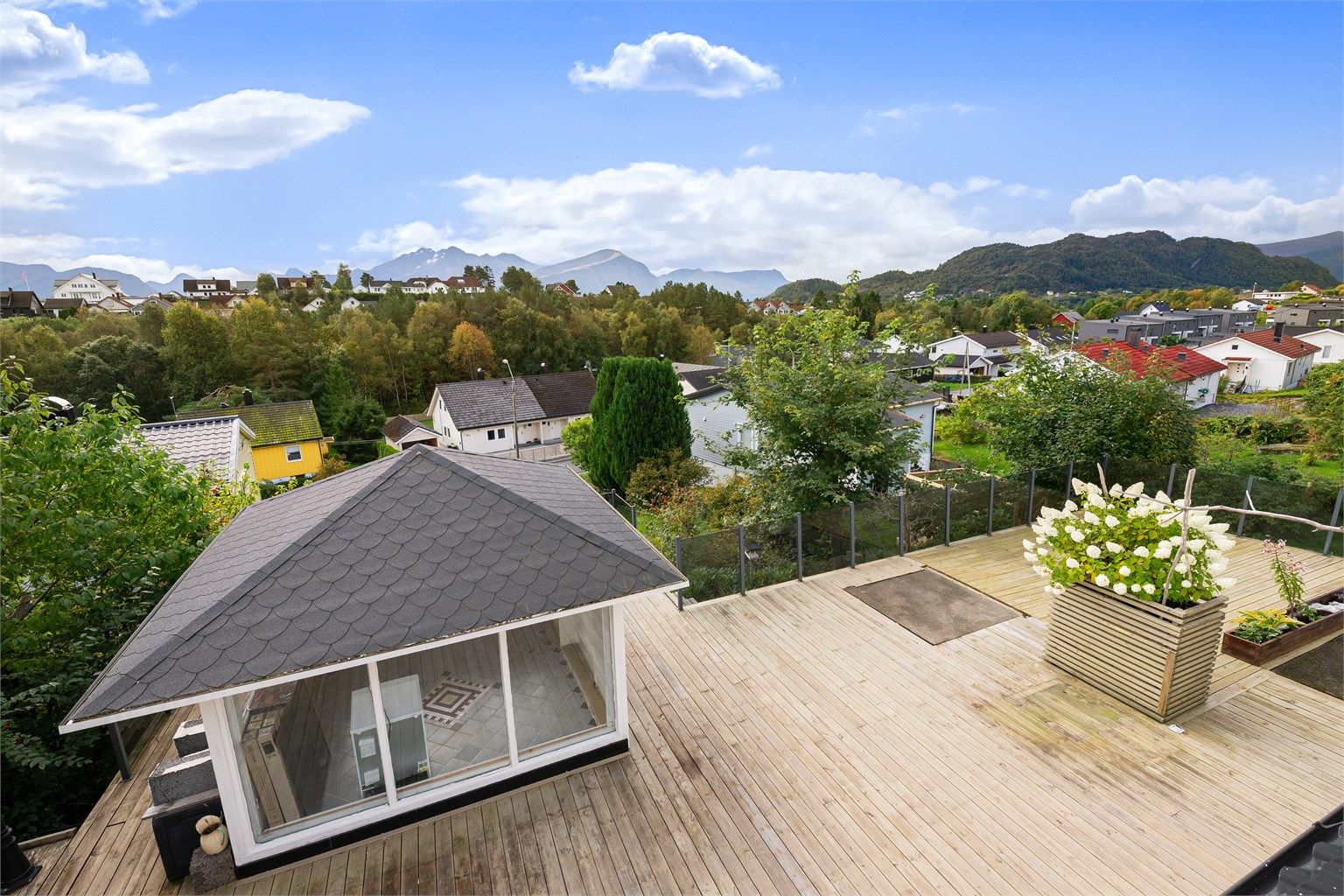 Utsikt fra balkongen i hovedetasjen mot stor terrasse på terreng med oppført lysthus
