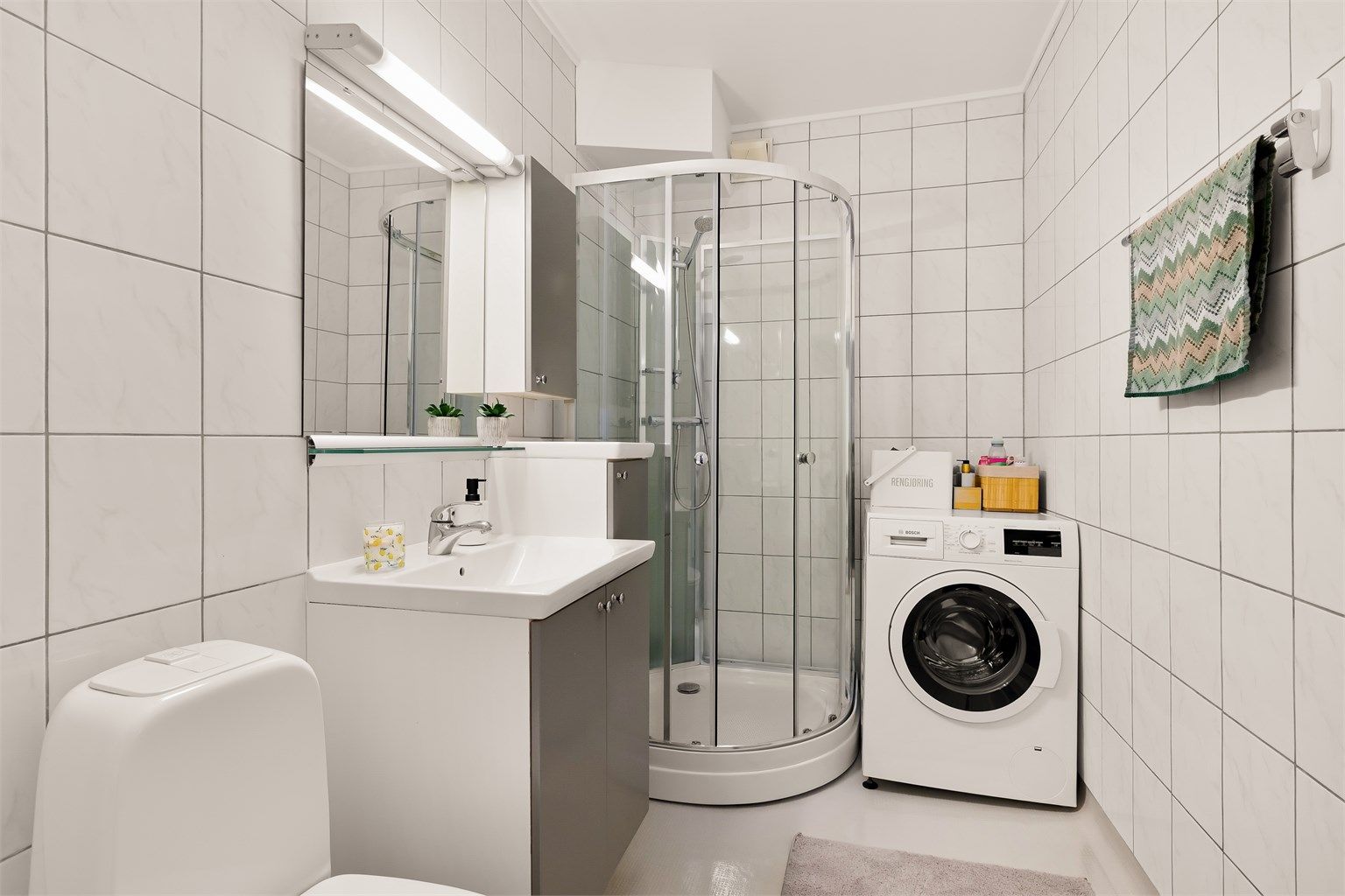 Badet har gulvbelegg og flislagte vegger. Det montert innredning med slette fronter, heldekkende servant. Badet er også utstyrt med overskap, speil, overlys og stikkontakt. Dusjkabinett og gulvstående toalett. Opplegg for vaskemaskin.