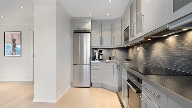 Integrert stekeovn og platetopp fra 2025. Frittstående oppvaskmaskin og kjøleskap med fryser. Ventilator i overskap. Det er lagt fliser mellom kjøkkenbenk og overskap.