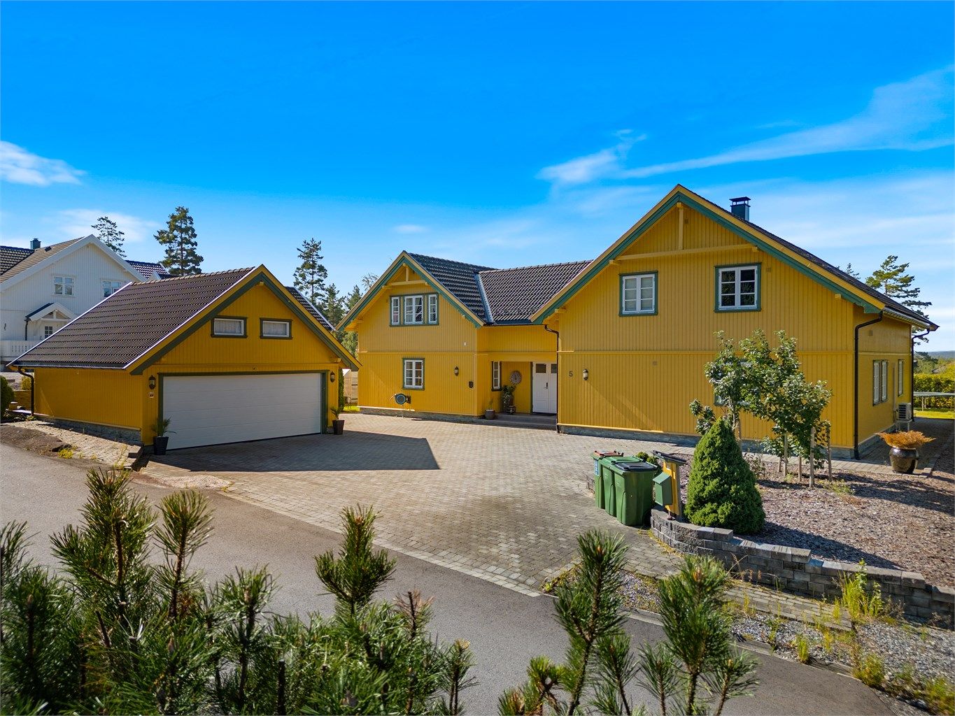 Foran boligen og garasjen møter du en elegant steinlagt gårdsplass som strekker seg videre rundt begge sider av huset, og binder eiendommen harmonisk sammen.