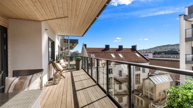 Balkongen er i betongkonstruksjon med rekkverk av metall og glass, og terrassebord på gulvet.
