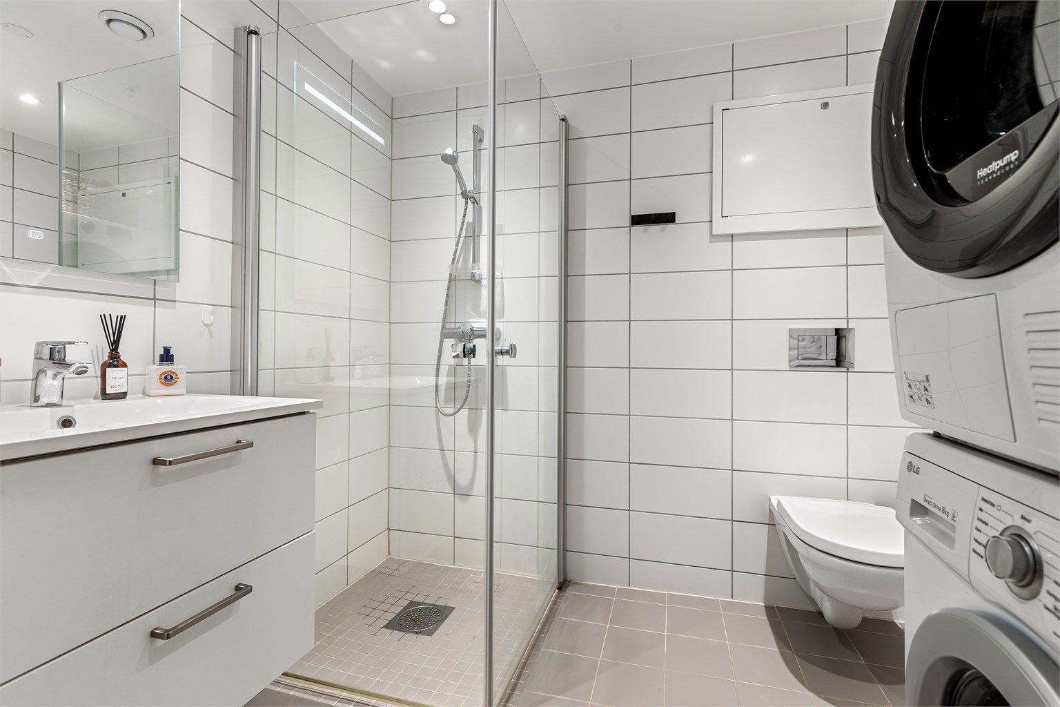 Innredet med dusjhjørne med glassdører, vegghengt toalett og vegghengt servantskap med speil og belysning. Opplegg for vaskemaskin og tørketrommel