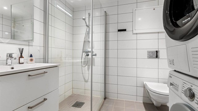 Innredet med dusjhjørne med glassdører, vegghengt toalett og vegghengt servantskap med speil og belysning. Opplegg for vaskemaskin og tørketrommel