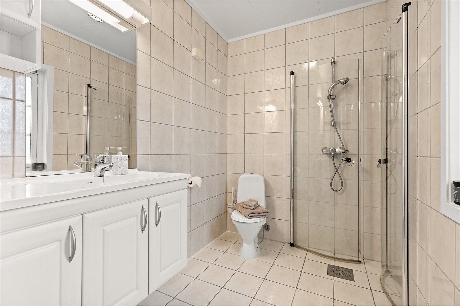 Bad med flislagt gulv, elektrisk gulvvarme og flislagte vegger. Vegghengt servantskap med heldekkende servant og ett-greps armatur.