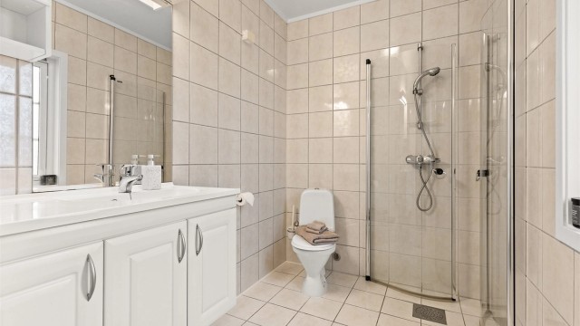 Bad med flislagt gulv, elektrisk gulvvarme og flislagte vegger. Vegghengt servantskap med heldekkende servant og ett-greps armatur.