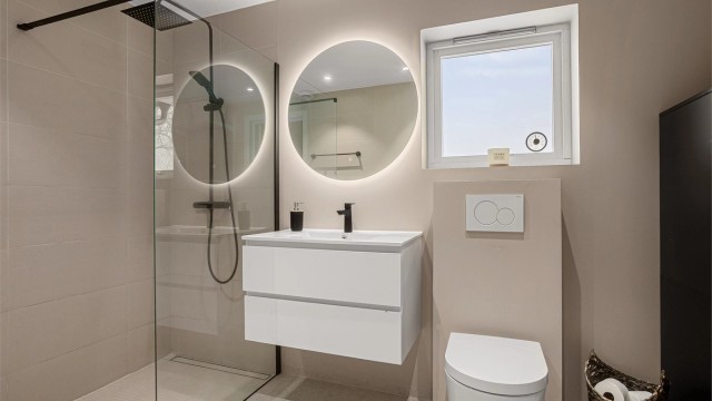 Innredet med dusjhjørne med skillevegg av glass, vegghengt toalett samt vegghengt servantskap med speil og integrert belysning