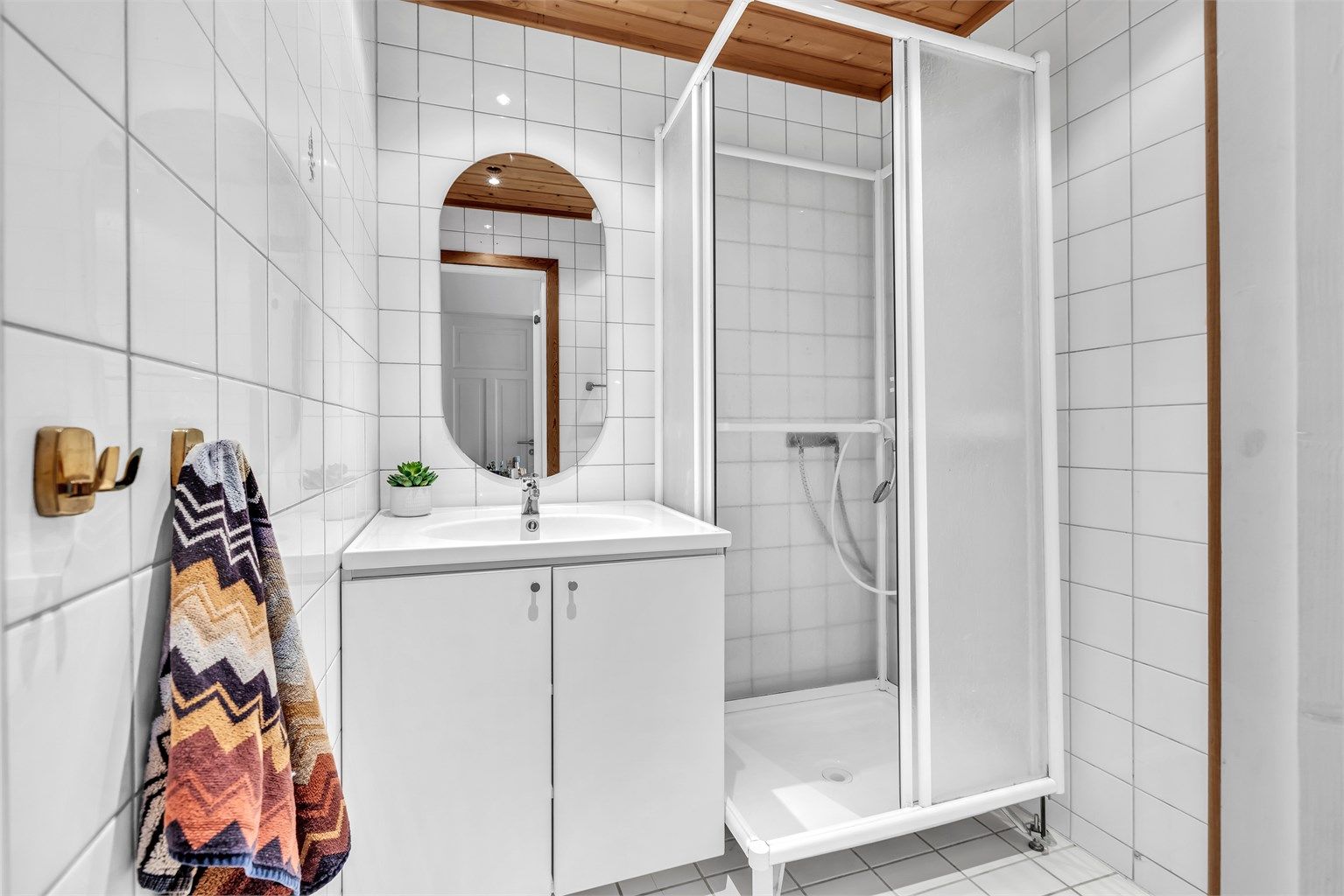 Flislagt baderom i første etasje. Vegghengt servantskap med glatte skapdører, servant, speil over servanten og et dusjkabinett.