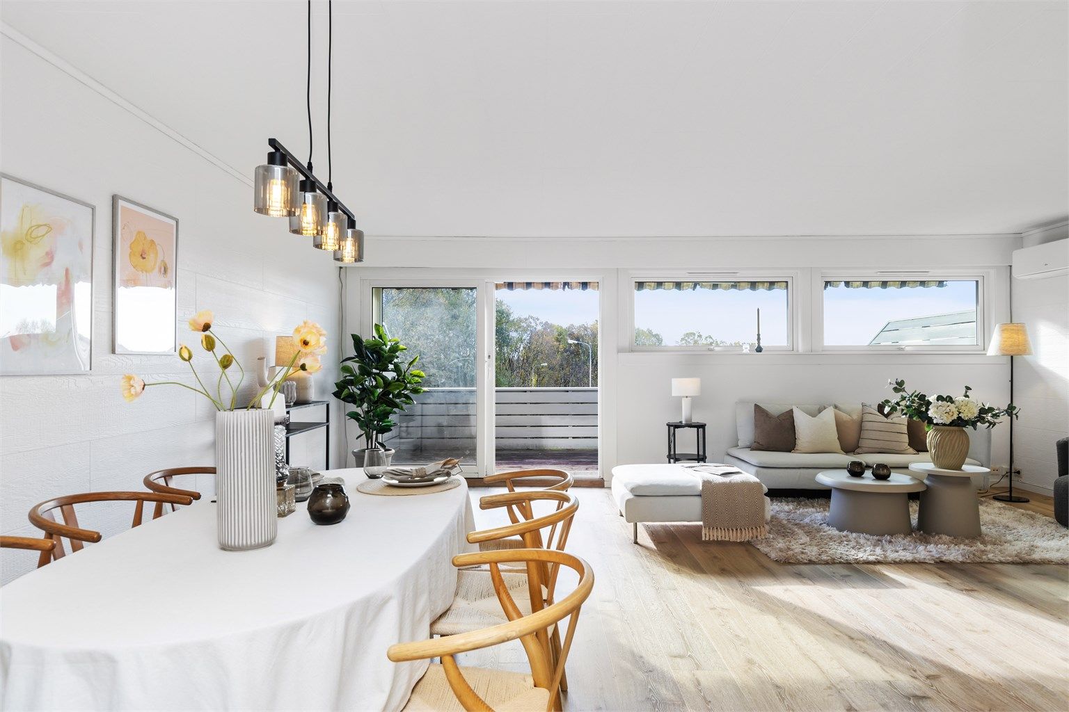 Lyst og moderne stue med åpen planløsning, store vinduer og utgang til balkong. Rommet er innredet med en elegant spiseplass, lyse tremøbler og en koselig sofakrok i nøytrale toner som skaper en harmonisk og innbydende atmosfære.