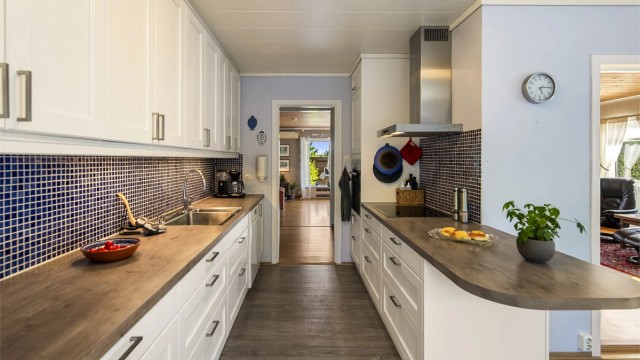 Kjøkkeninnredningen har hvite, profilerte fronter og benkeplater i laminat med nedfelt oppvaskkum. Fliser mellom overskap og benkeplass. Kjøkkenet har rikelig med skap- og benkeplass.
