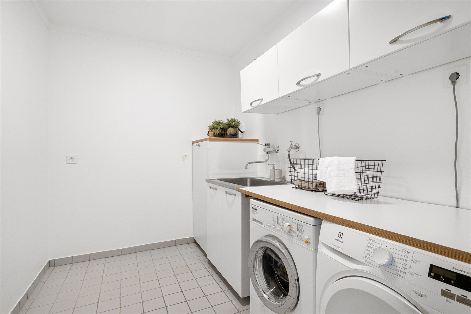 Meget praktisk og romslig vaskerom med rikelig plass til oppbevaring i skap på vegg.Opplegg for vaskemaskin og tørketrommel, som er plassert ved siden av hverandre og med benkeplate over. Utslagsvask. Det er fliser på gulv og slette vegger. 