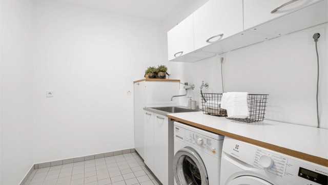 Meget praktisk og romslig vaskerom med rikelig plass til oppbevaring i skap på vegg.Opplegg for vaskemaskin og tørketrommel, som er plassert ved siden av hverandre og med benkeplate over. Utslagsvask. Det er fliser på gulv og slette vegger.