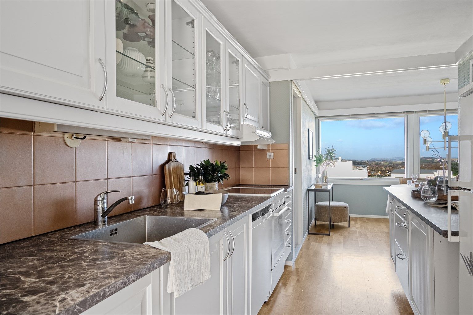 Kjøkkeninnredning i lyse farger og med grå/hvit benkeplate. Det er rikelig med oppbevaring i skuffer og skap, samt gode arbeidsflater på benk. Fin utsikt mot Stokka. Malte fliser på vegg over benk.