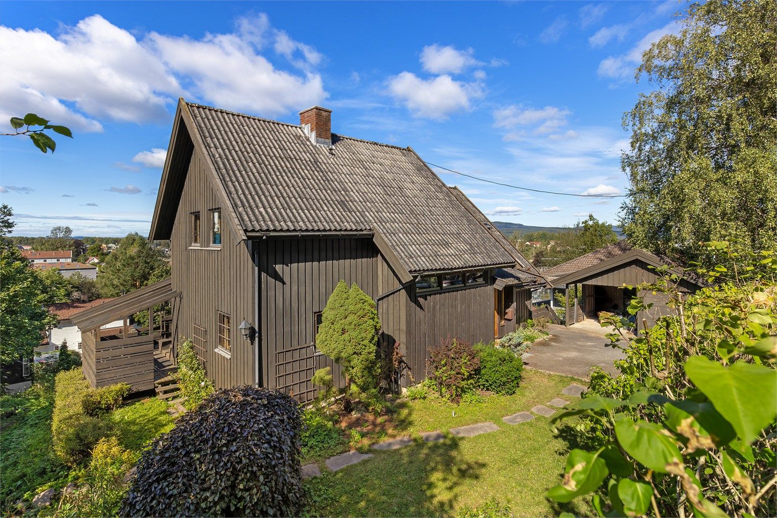 Fasaden sett fra Kurveien. Asfaltert gårdplass, hellelagt gangsti rundt huset til hagesiden. Garasje/carport med lagringsrom under