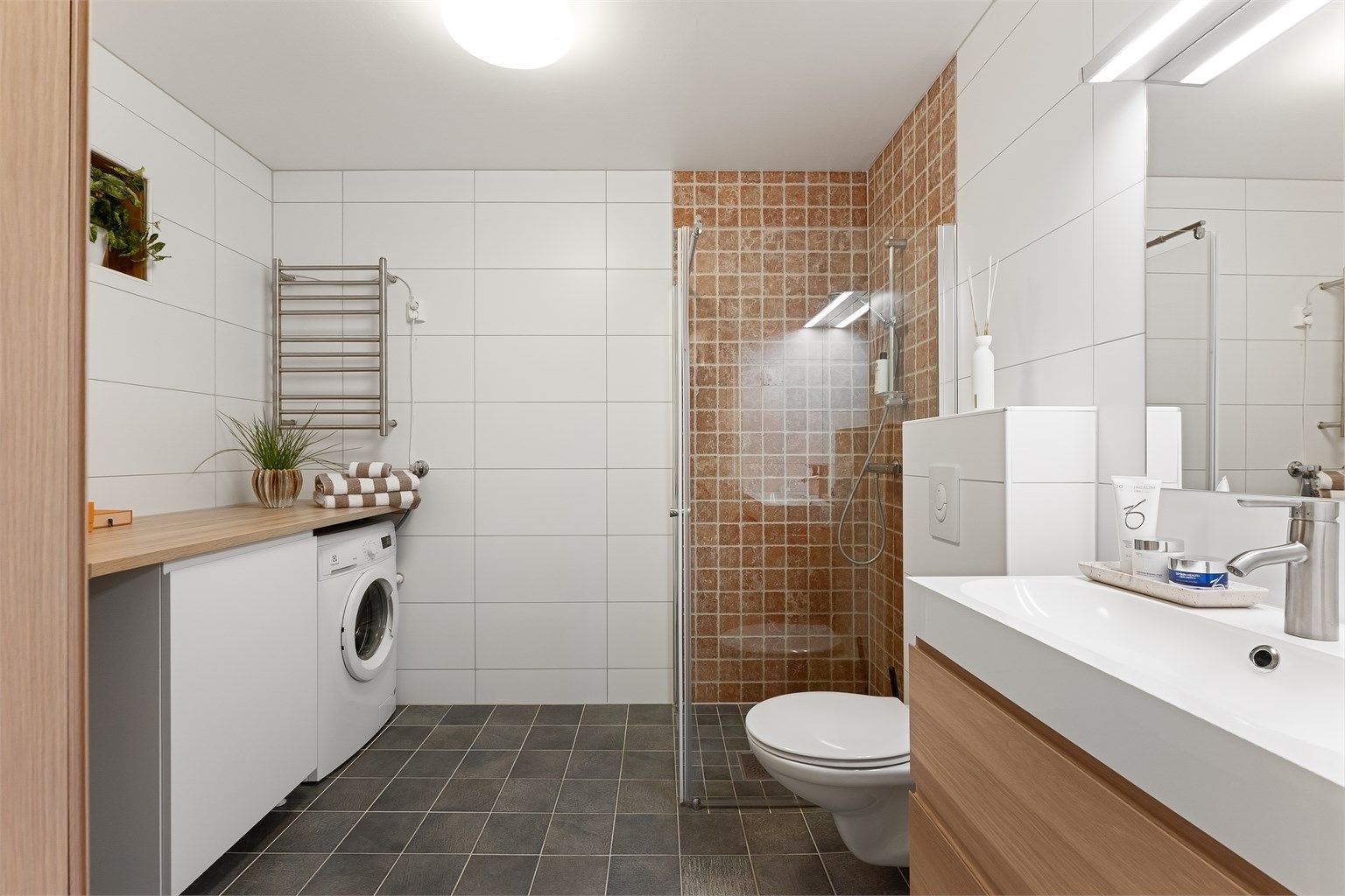 Pent baderom med fliser på gulv med gulvvarme, fliser på vegger og malt himling. Badet har vegghengt toalett, vegghengt servantinnredning, speil med overlys og dusjhjørne med dører