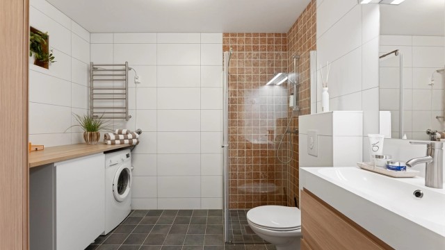 Pent baderom med fliser på gulv med gulvvarme, fliser på vegger og malt himling. Badet har vegghengt toalett, vegghengt servantinnredning, speil med overlys og dusjhjørne med dører