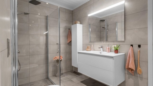 Badet er fra byggeår og har flislagt gulv med gulvvarme samt flislagte vegger. Innredningen består av vegghengt servantinnredning med ovenpåliggende servant og armatur, speil med overlys og stikkontakt samt vegghengt høyskap.