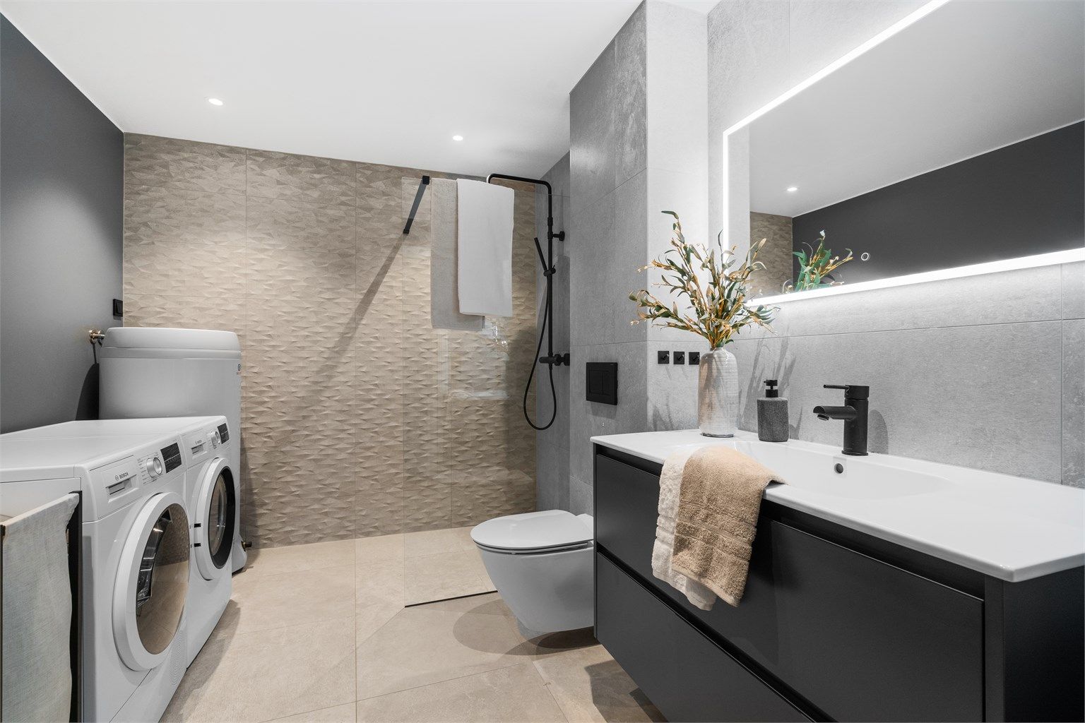 Varmekabler i gulv, vegghengt toalett, stor dusj.  Opplegg for vaskemaskin samt plass til tørketrommel. Grå fliser på gulv og vegg, glassvegg i dusj. 
