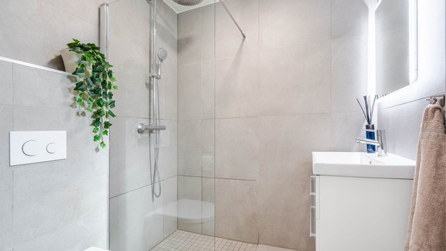 Badet har veggmontert toalett, servant med skap og et nedsenket dusjhjørne med glassvegg.
