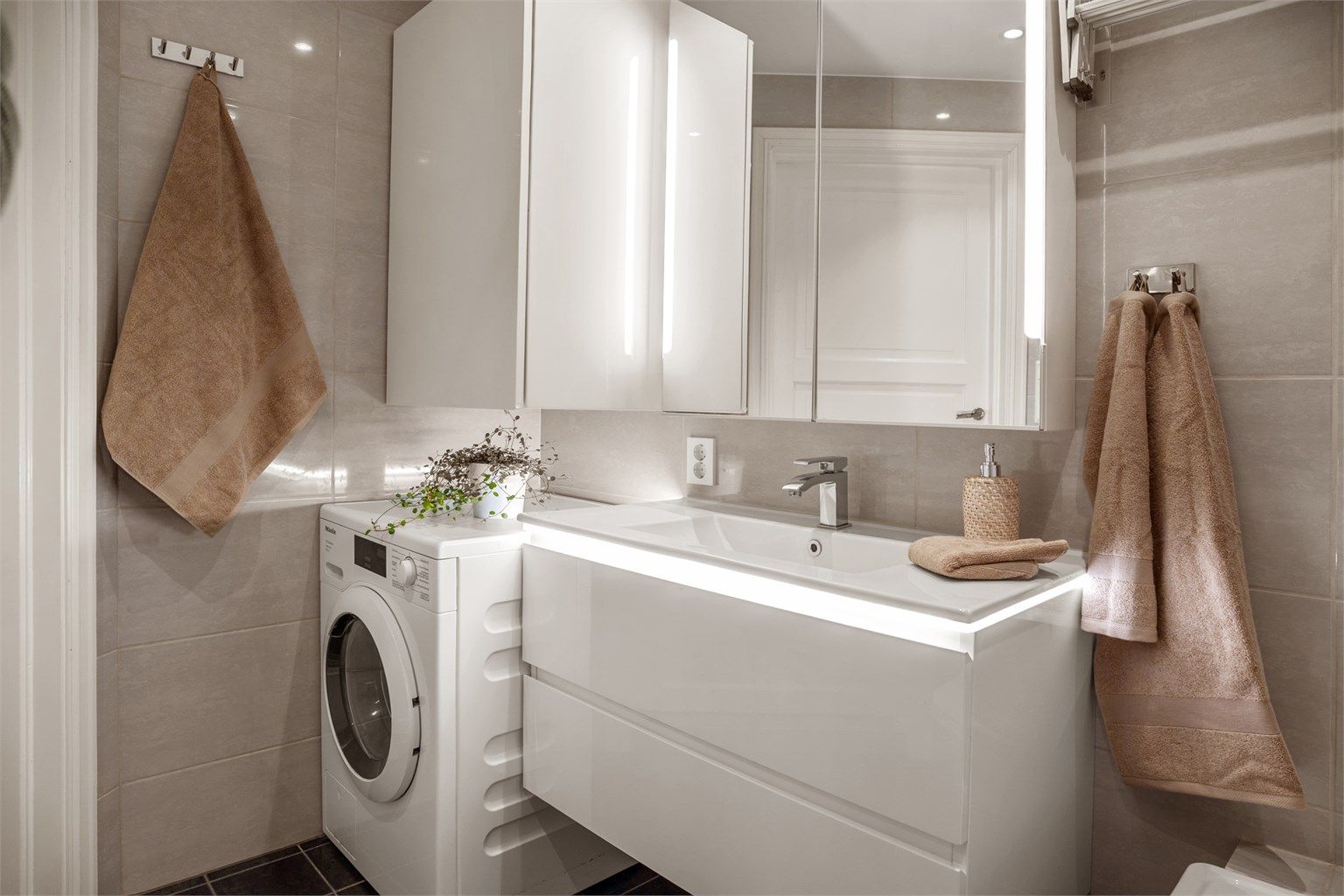 Badet er utstyrt med vegghengt servant, speilskap med belysning og opplegg for vaskemaskin med praktisk overskap til oppbevaring.