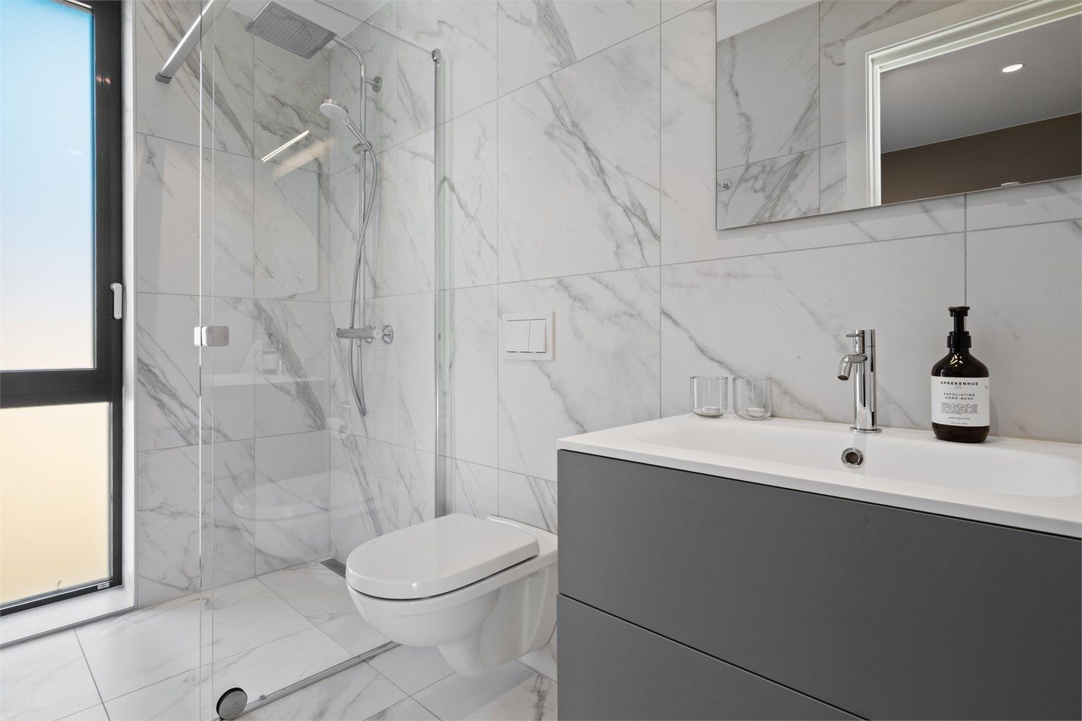 Badet har flislagte gulv og vegger, innredet med servant, vegghengt toalett og dusjhjørne. 