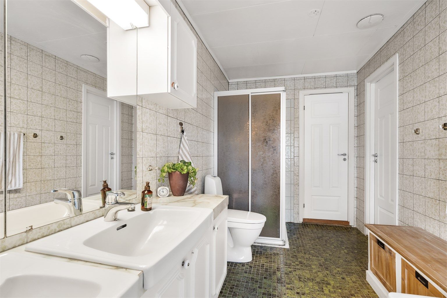 Badet er innredet med fliser på både gulv og vegger. Gulvvarme og folierte himlingsplater. Vegghengt servantinnredning med overskap og to servanter. Dusjnisje med skyvedører og badekar