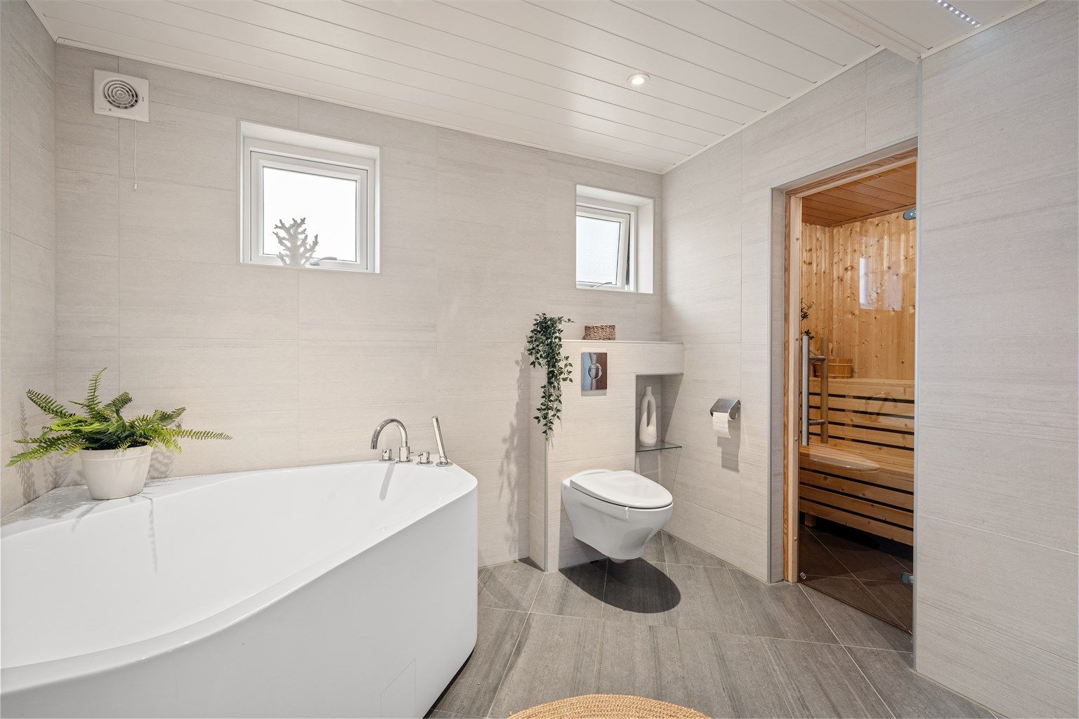 Badet gir en god romfølelse med delikate fargevalg og er innredet med vegghengt toalett, badekar og dusjnisje. De små vinduene gir godt med naturlig lys uten innsyn.