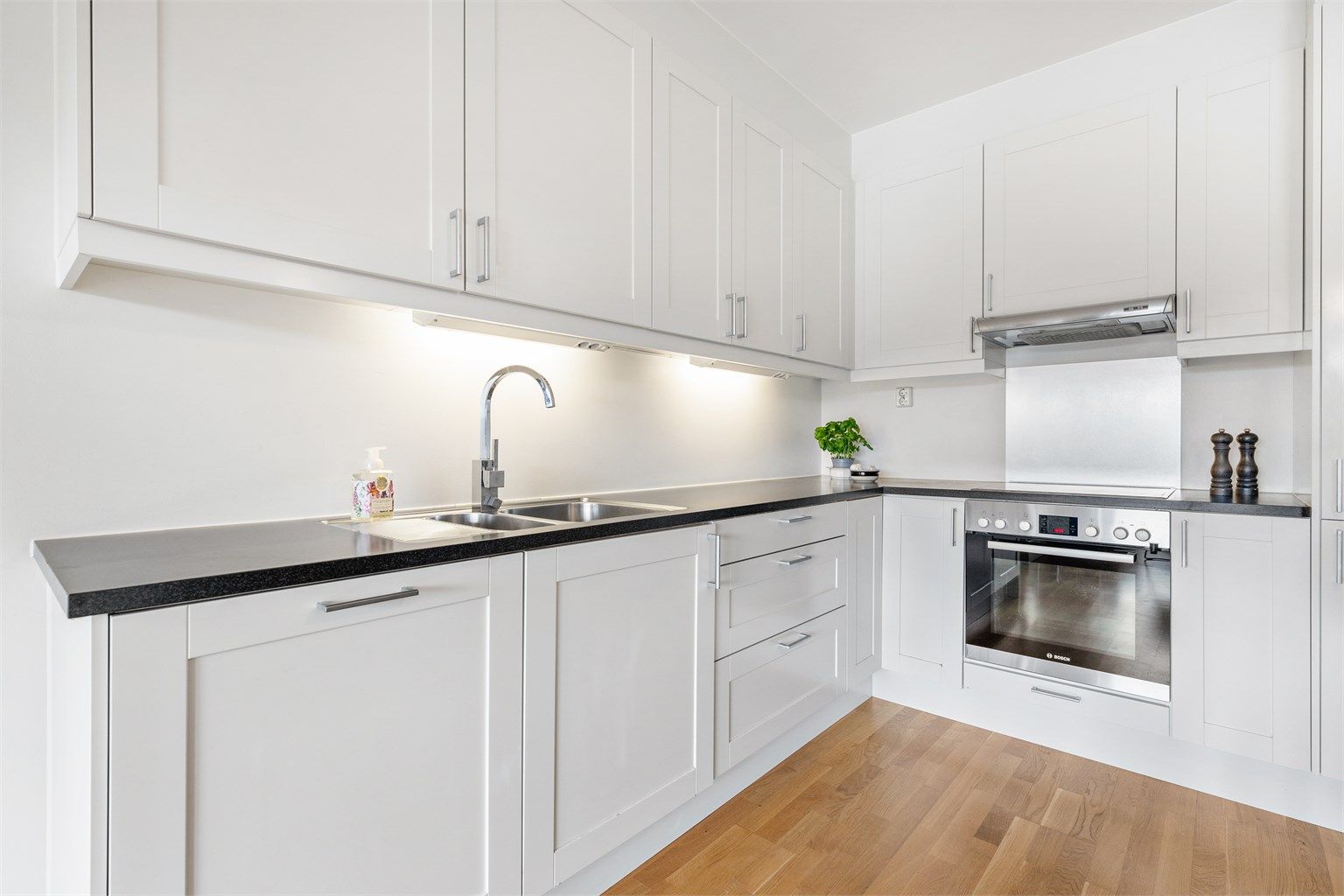 Malt slett flate mellom kjøkkenbenk og overskap, stenkeplate bak nedfelt keramisk platetopp. Stikkontakt på vegg. Benkeskapsbelysning og stikkontakter under overskap. Komfyr og oppvaskmaskin under benk. Kjøleskap med frysedel i høyskap.