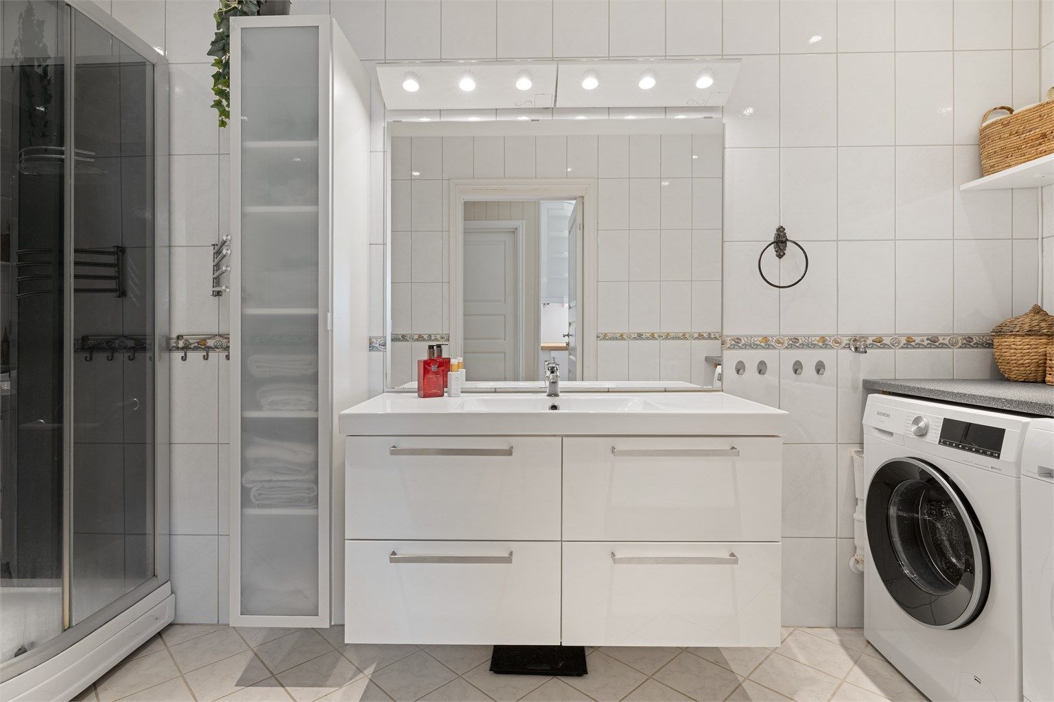 Badet er flislagt og innredet med servant, speil med overlys og stikkontakt over servanten. Videre er badet utstyrt med vegghengt høyskap, dusjkabinett og gulvstående toalett.
