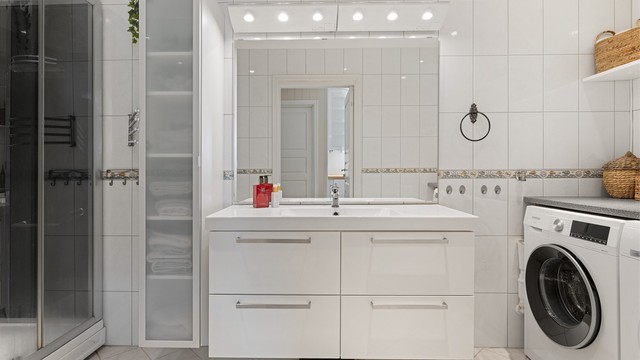 Badet er flislagt og innredet med servant, speil med overlys og stikkontakt over servanten. Videre er badet utstyrt med vegghengt høyskap, dusjkabinett og gulvstående toalett.